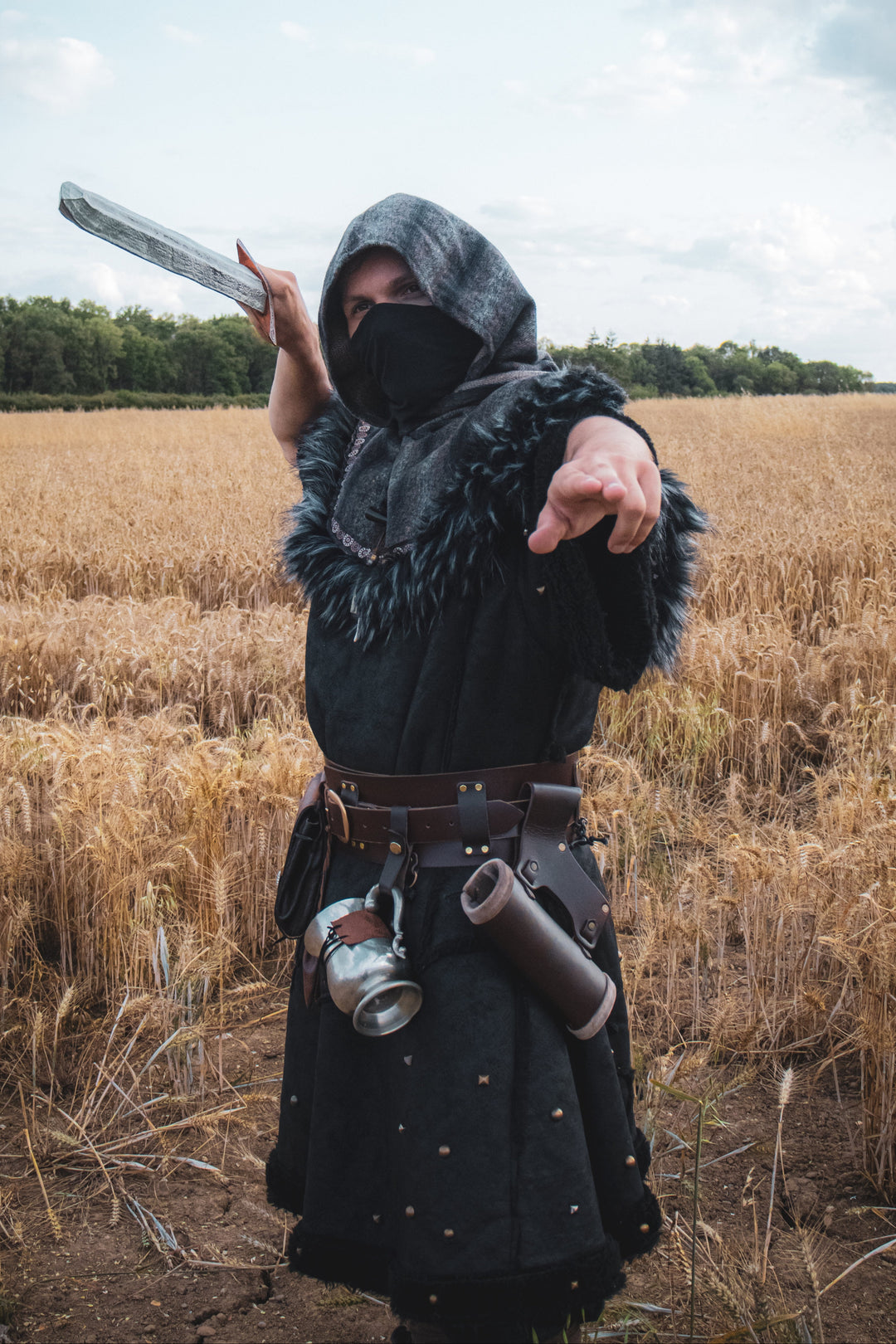 Person in a hooded cloak holding a weapon in a field.