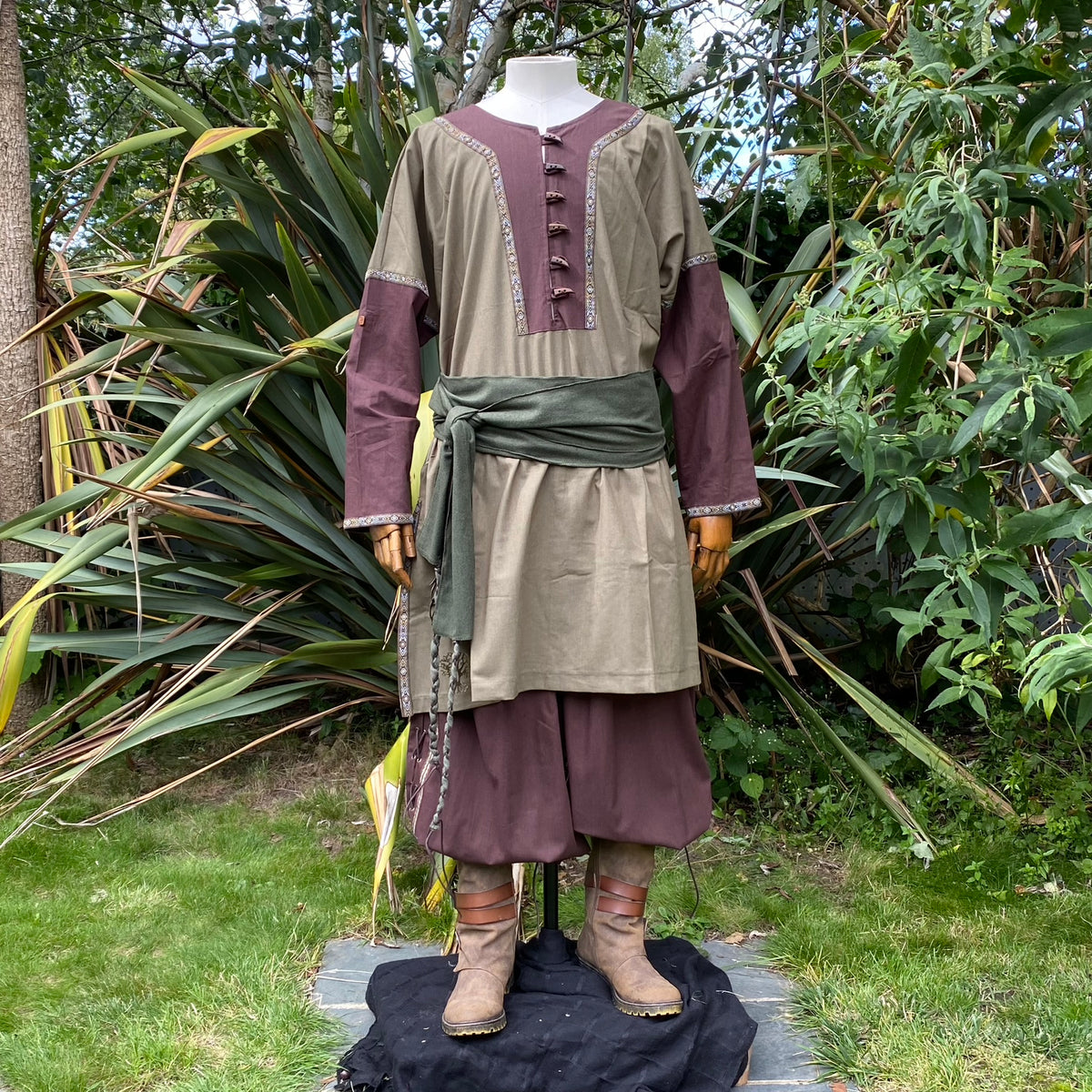 Medieval LARP outfit on mannequin, green sash, front view with lush greenery.