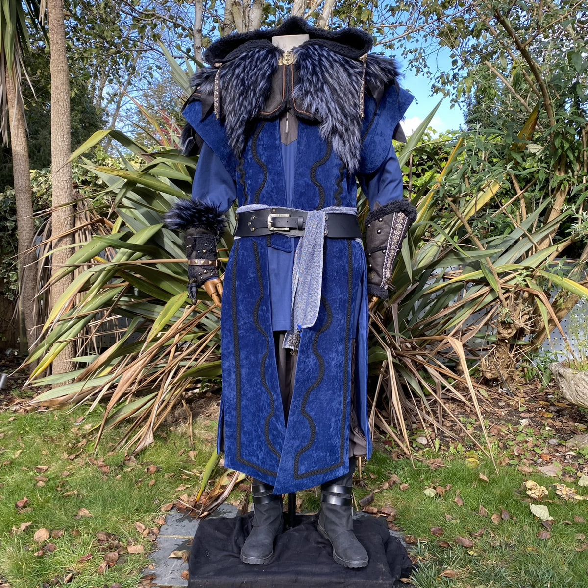 Blue medieval LARP costume with fur trim, greenery backdrop, front view.