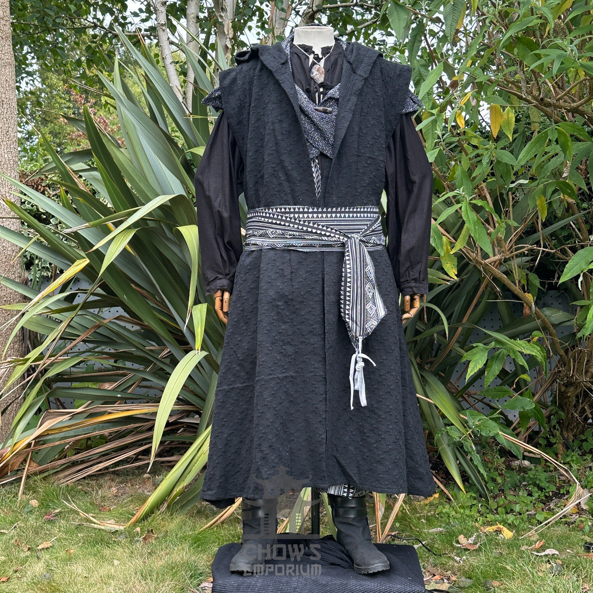 Medieval LARP attire in a garden, hooded waistcoat, detailed sash, boots.