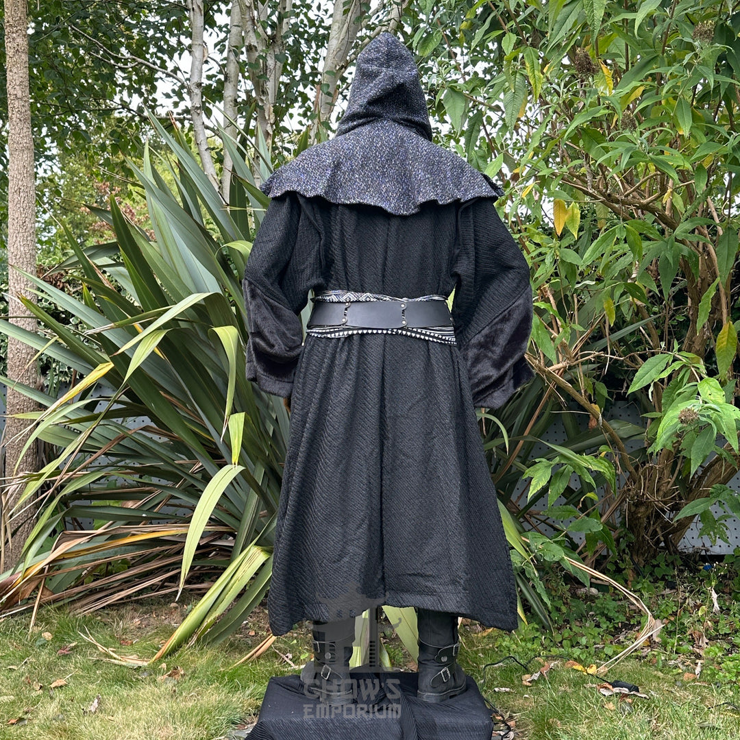 Black LARP outfit with ornate hood, belt, and sash, rear view amidst greenery.