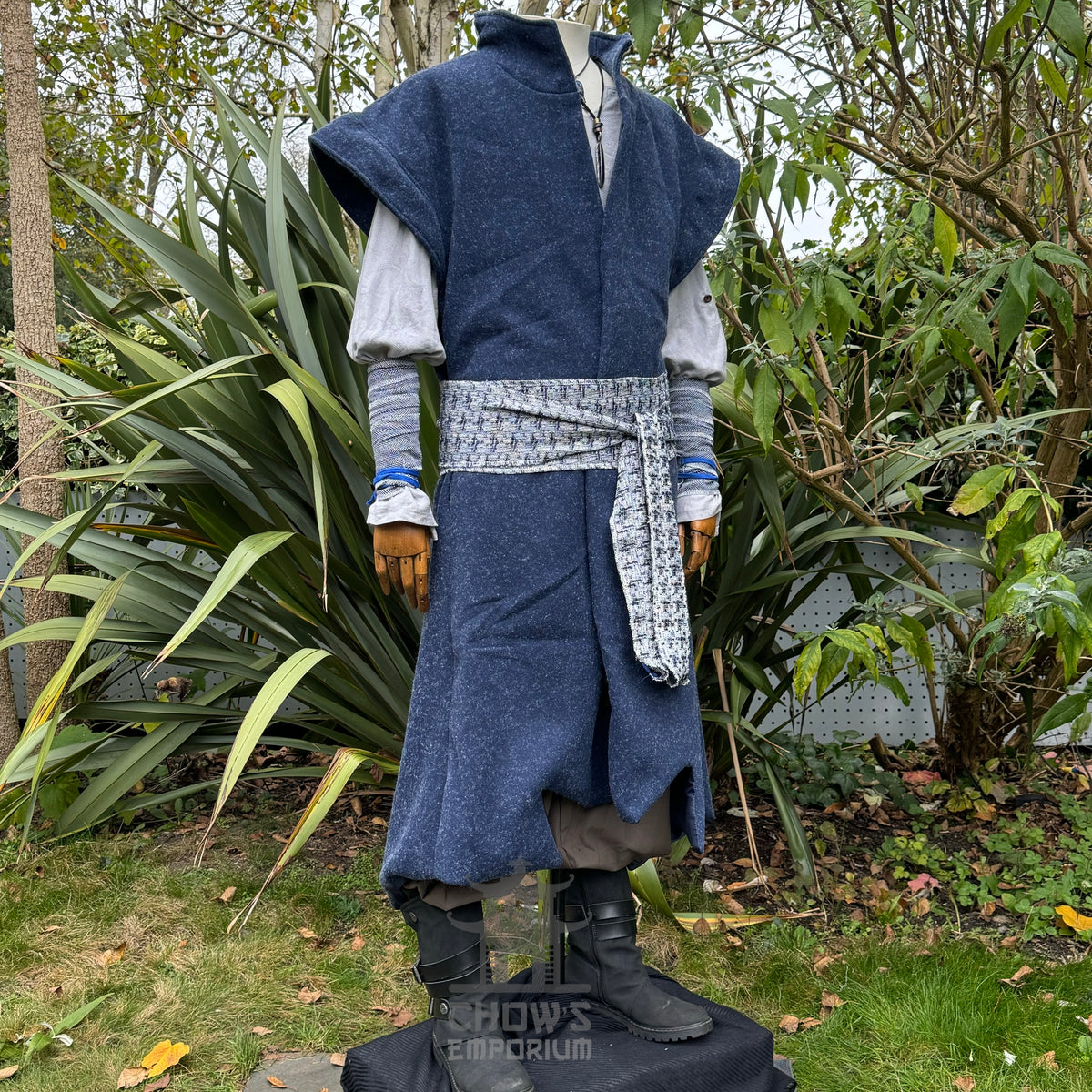 Medieval LARP costume side view, featuring a blue waistcoat, tunic, trousers, arm wraps, sash.
