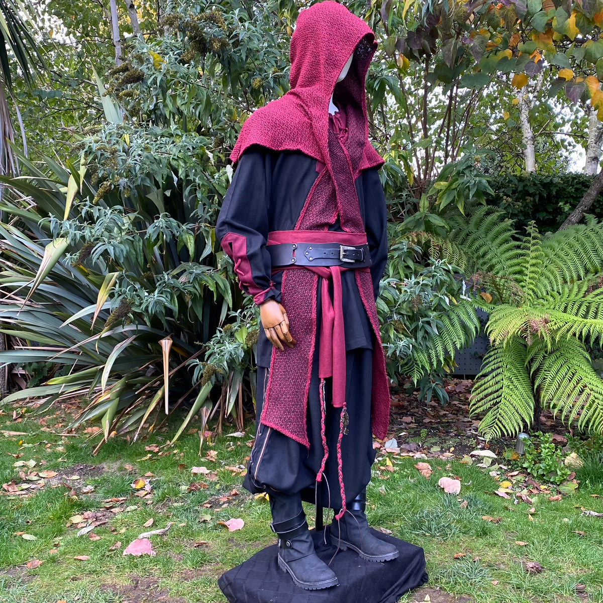 Red woolen hood draped over mannequin amidst lush garden, medieval LARP attire.