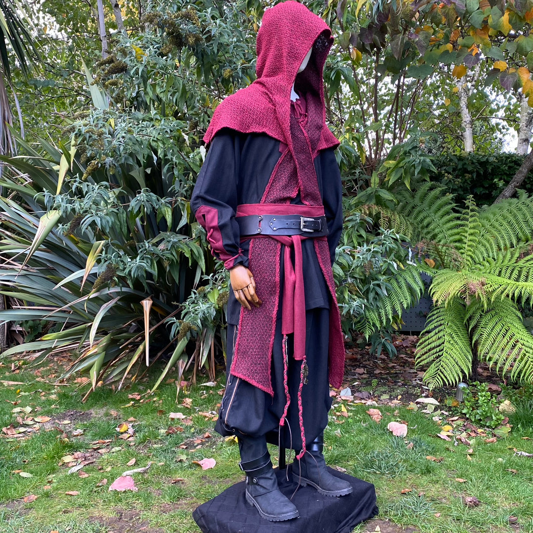 Red woolen hood draped over mannequin amidst lush garden, medieval LARP attire.