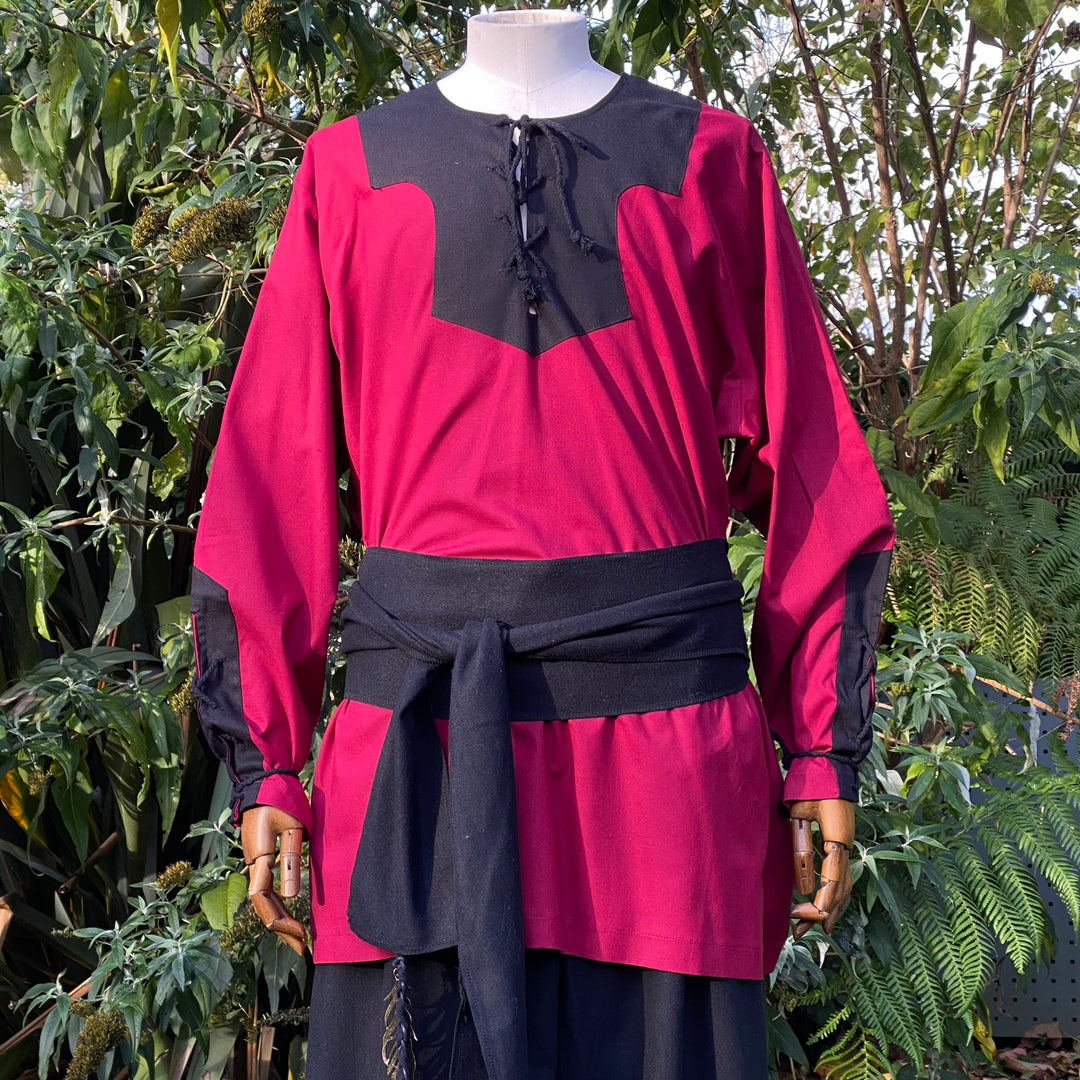 Mannequin in red-black medieval outfit with sash, surrounded by greenery.
