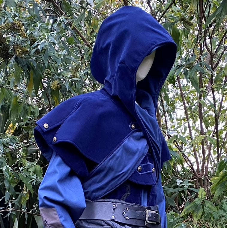 Cloaked figure in blue medieval LARP hooded outfit, side forest view.