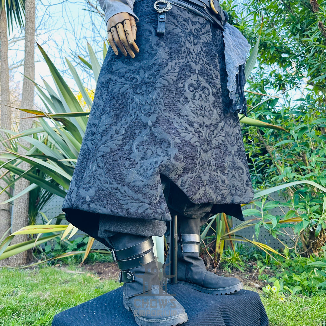 Close-up of the black brocade waistcoat hem with a V-cut tailcoat front, worn over grey trousers and leather boots