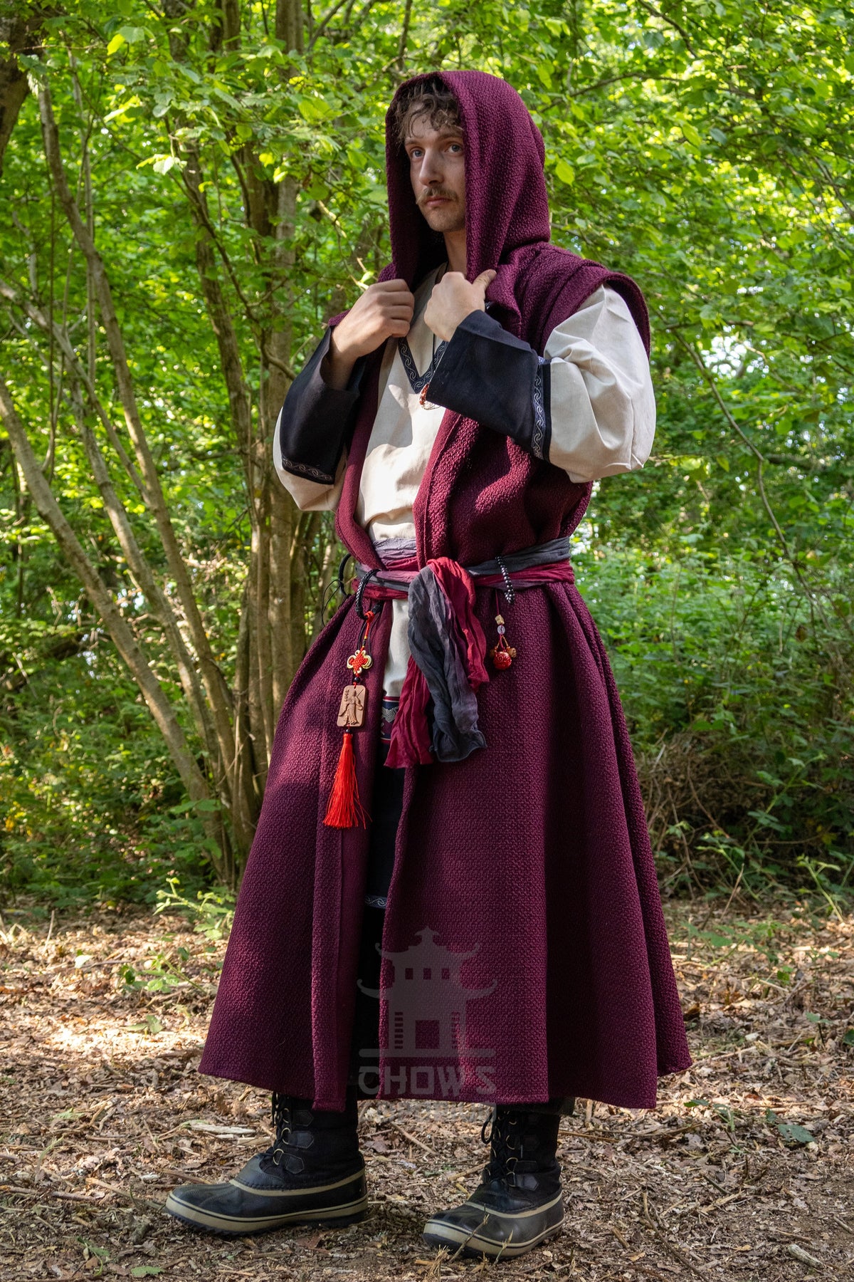 A man wearing a medieval-style costume with a deep burgundy hooded tunic, white sleeves, and a dark belt stands outdoors in a wooded setting. The image has a visible 'CHOWS' watermark near the bottom.