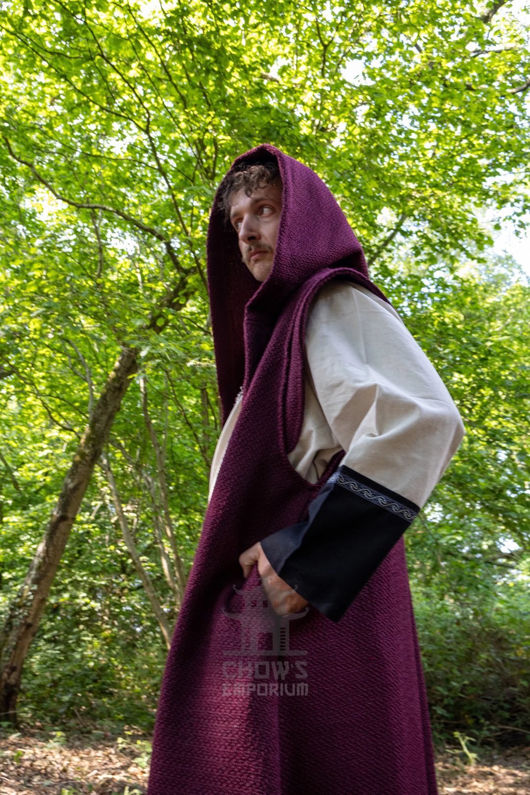 A man wearing a medieval-style long burgundy hooded cloak over a cream tunic and black sleeve, standing in a lush green forest. A watermark for &
