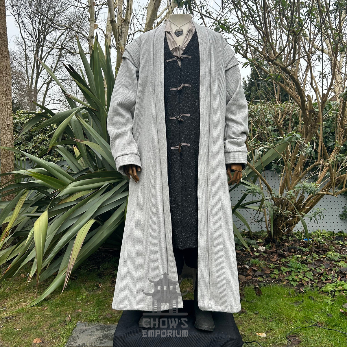 A light grey full-length robe displayed on a mannequin, featuring a herringbone pattern, in a garden setting.