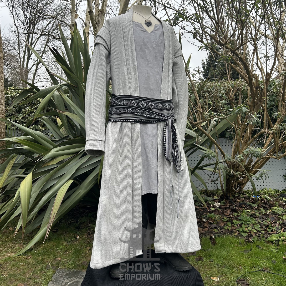 A light grey full-length robe displayed open with sash belt on a mannequin, shown from side view, featuring a herringbone pattern, in a garden setting.