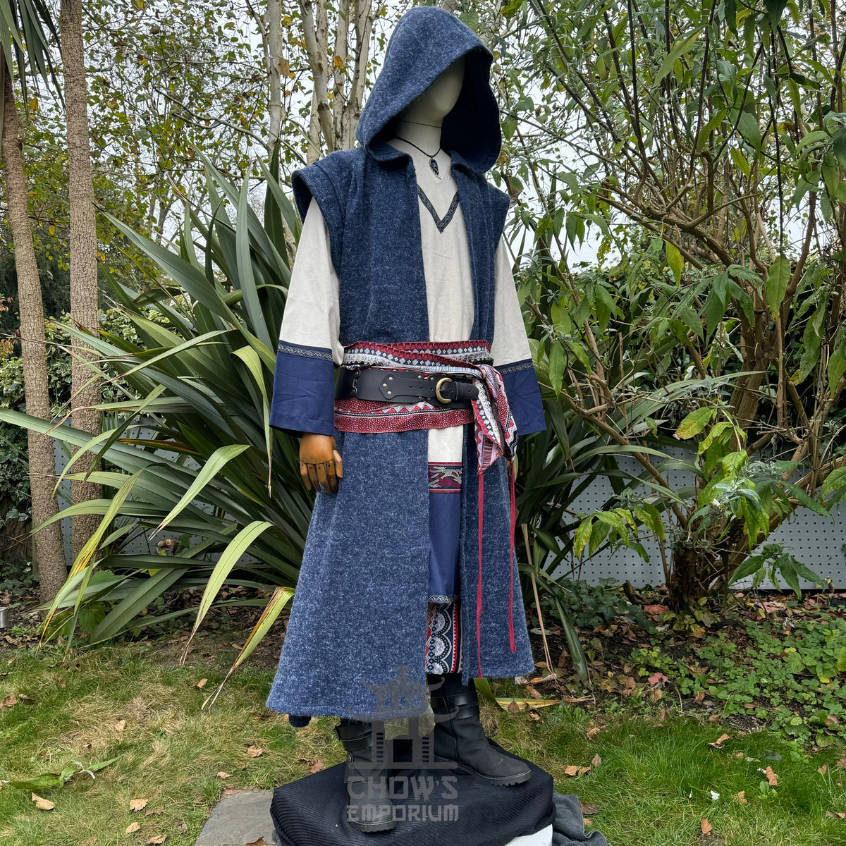 Mannequin wearing a blue hooded sleeveless robe with a patterned belt shown from side view in an outdoor setting.