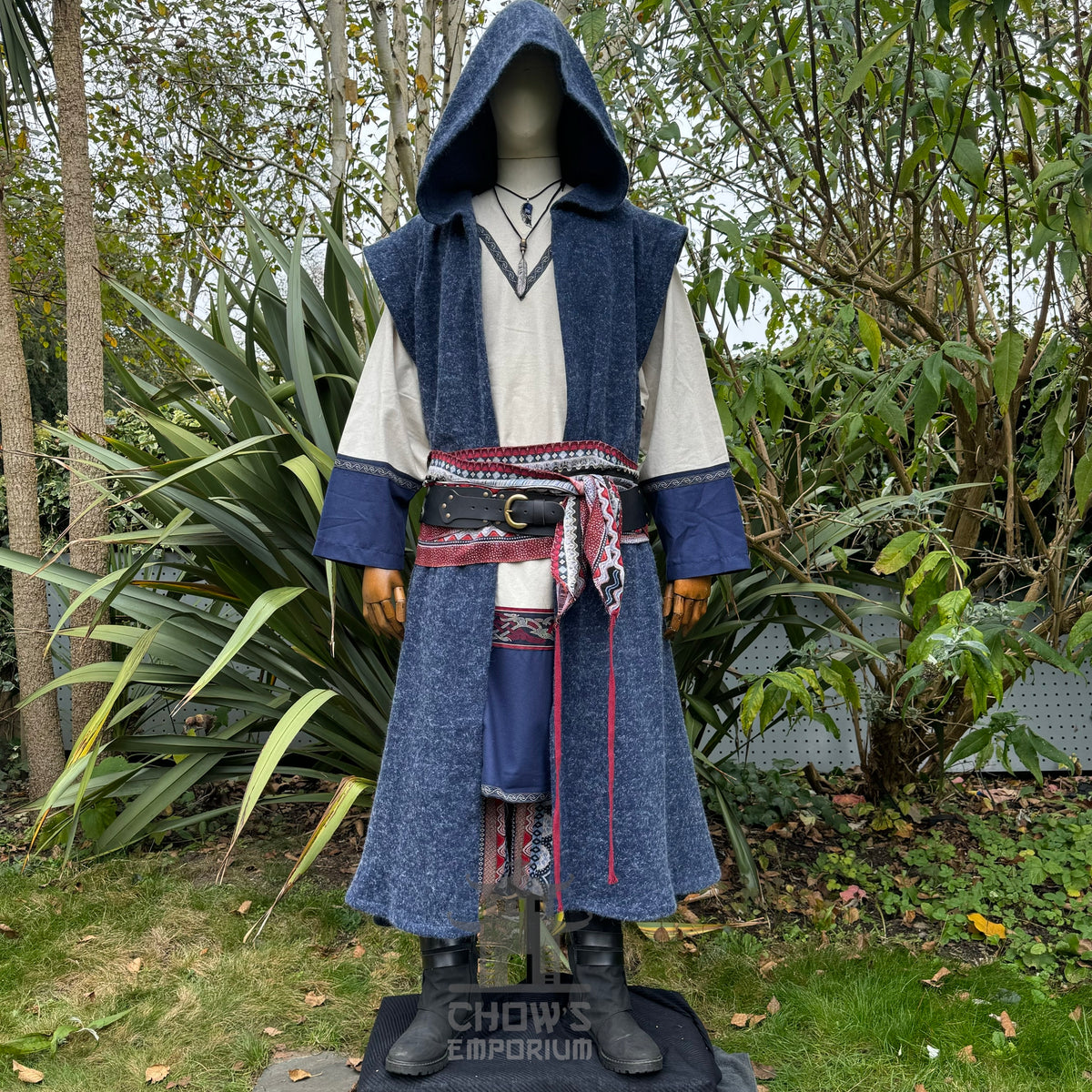 Mannequin wearing a blue hooded sleeveless robe with a patterned sash in an outdoor setting.