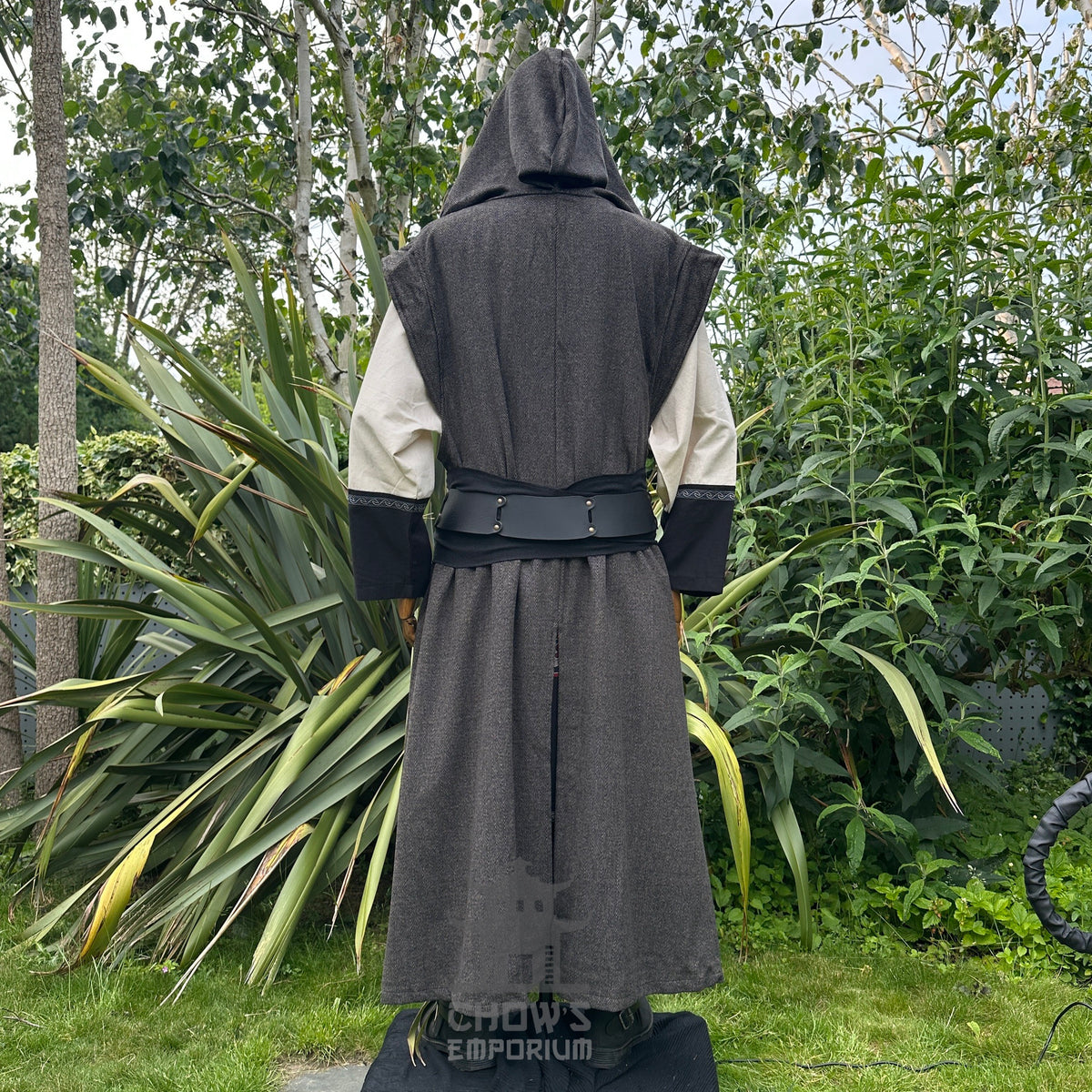 Back view of mannequin wearing hooded brown herringbone sleeveless robe with black belt, in garden setting.