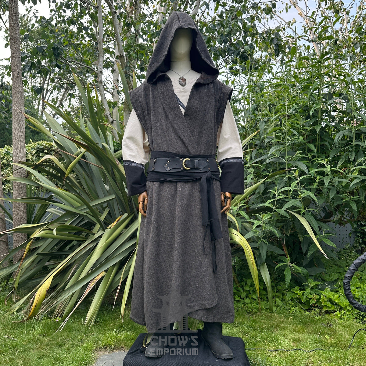 Mannequin in hooded brown herringbone sleeveless robe with black belt, in garden setting.