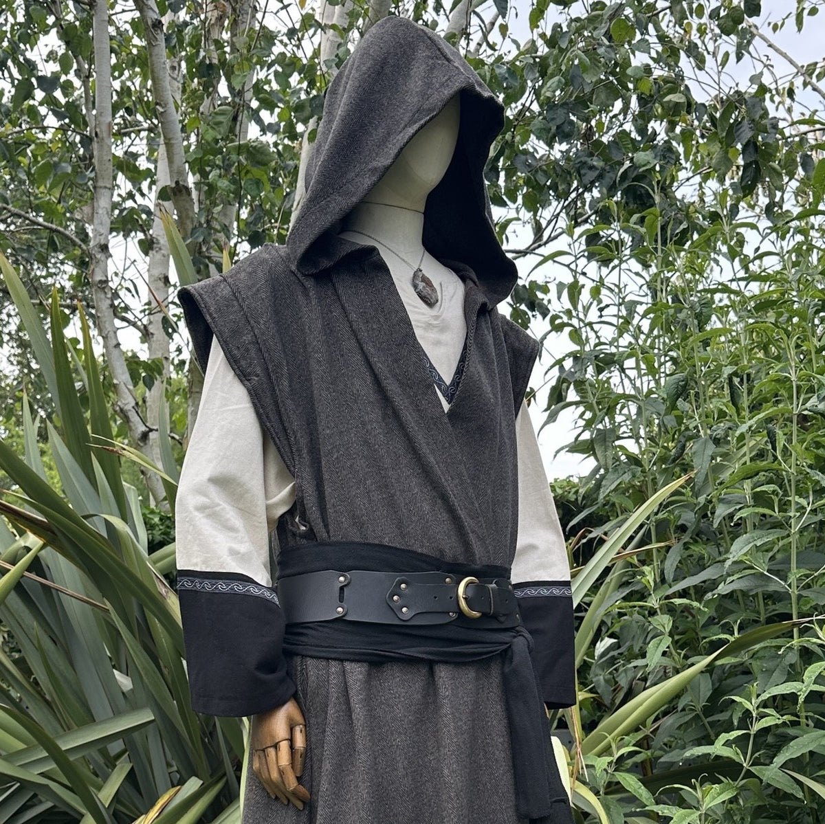 Cropped side view of mannequin in hooded brown herringbone sleeveless robe with black belt, in garden setting.