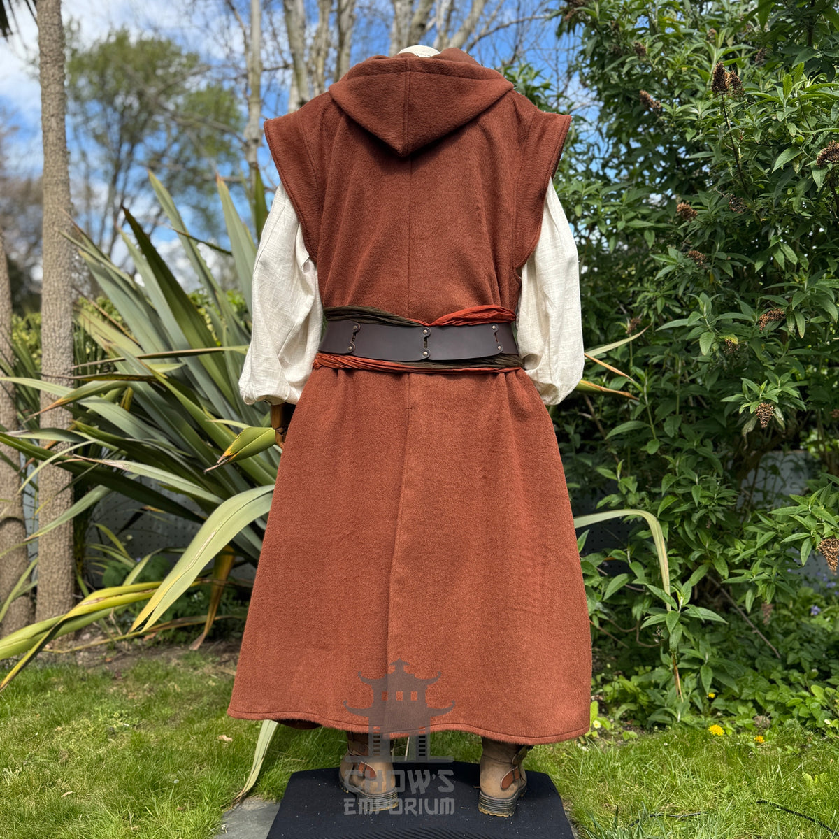 Mannequin wearing a medieval-style long rust robe, belted at the waist over a light beige shirt, shown from back view, standing outdoors in a garden with trees in the background. 