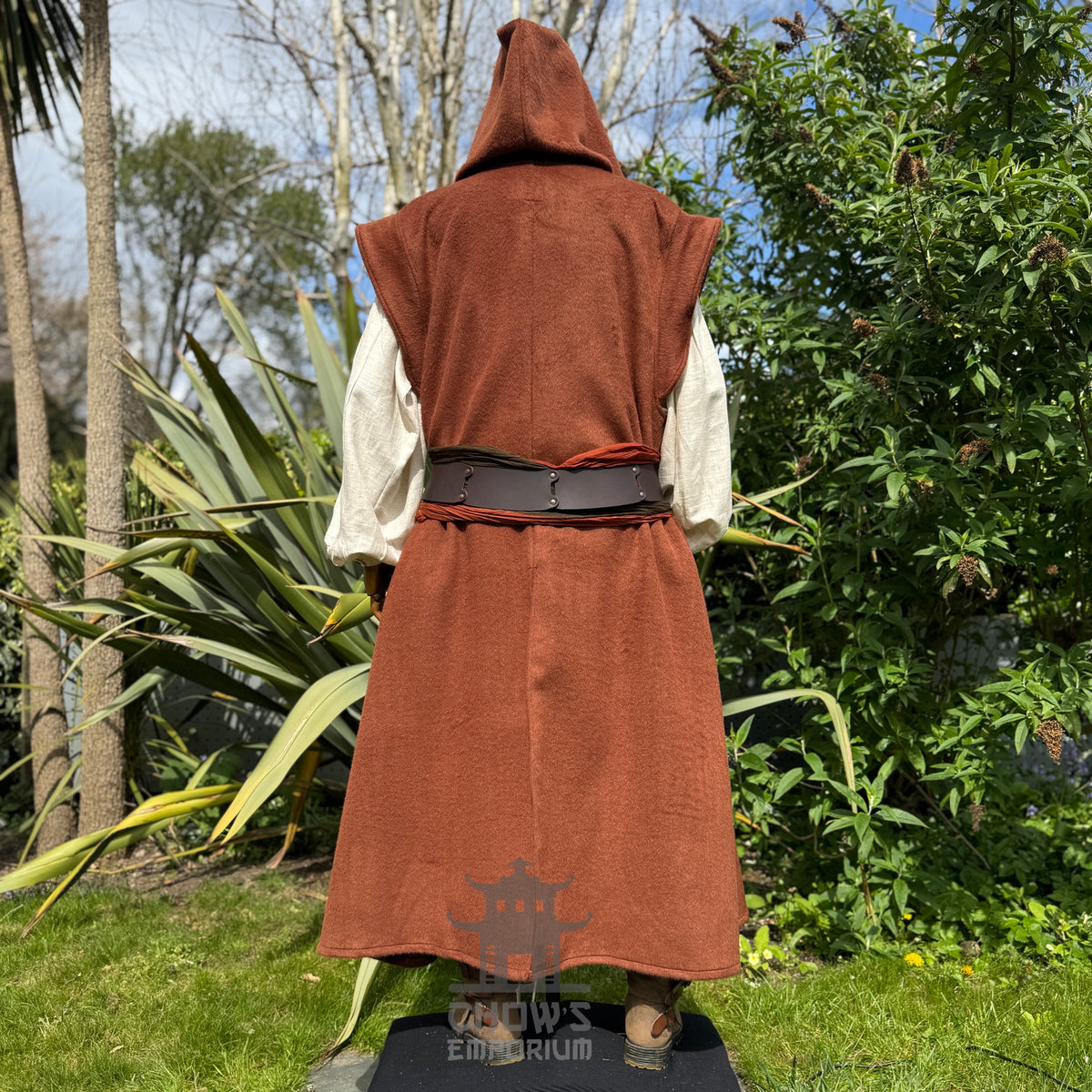 A mannequin wearing a medieval-style long rust hooded robe, shown full length from back view, standing outdoors in a garden with trees in the background. 