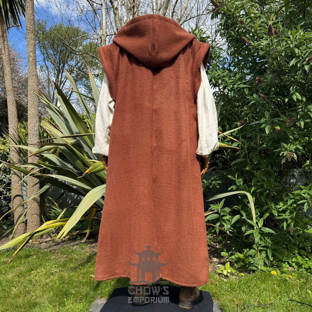 A mannequin wearing a medieval-style long rust hooded robe over a light beige shirt shown from back view, standing outdoors in a garden with trees in the background. 