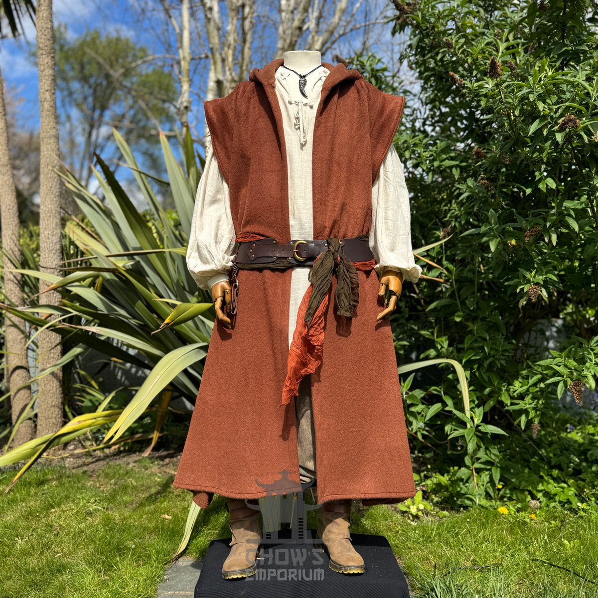 A mannequin wearing a medieval-style long rust hooded robe over a light beige shirt and brown trousers, belted at the waist, standing outdoors in a garden with trees in the background. 