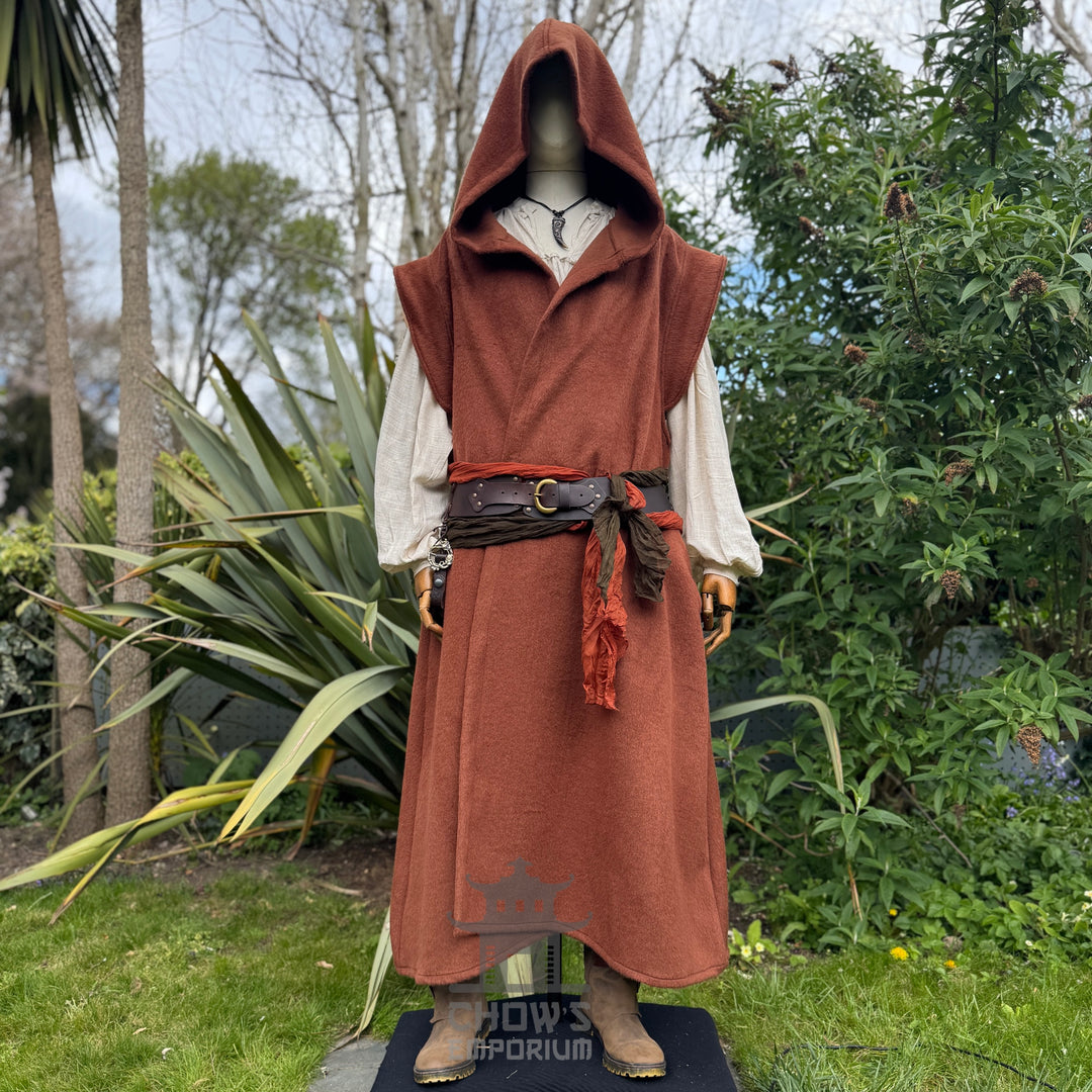A mannequin wearing a medieval-style long rust robe with hood up worn over a light beige shirt, belted at the waist, standing outdoors in a garden with trees in the background.