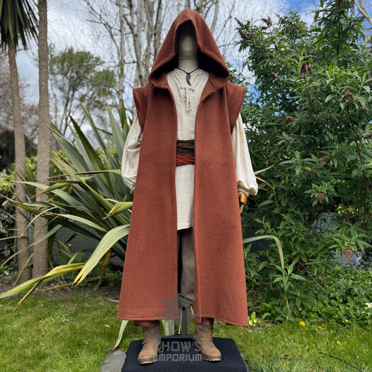 A mannequin wearing a medieval-style long rust hooded robe over a light beige shirt and brown trousers, standing outdoors in a garden with trees in the background. 