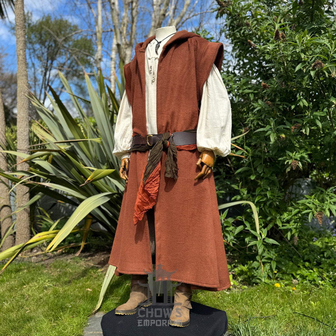 Mannequin wearing a medieval-style long rust robe, belted at the waist over a light beige shirt, shown from front side view, standing outdoors in a garden with trees in the background. 