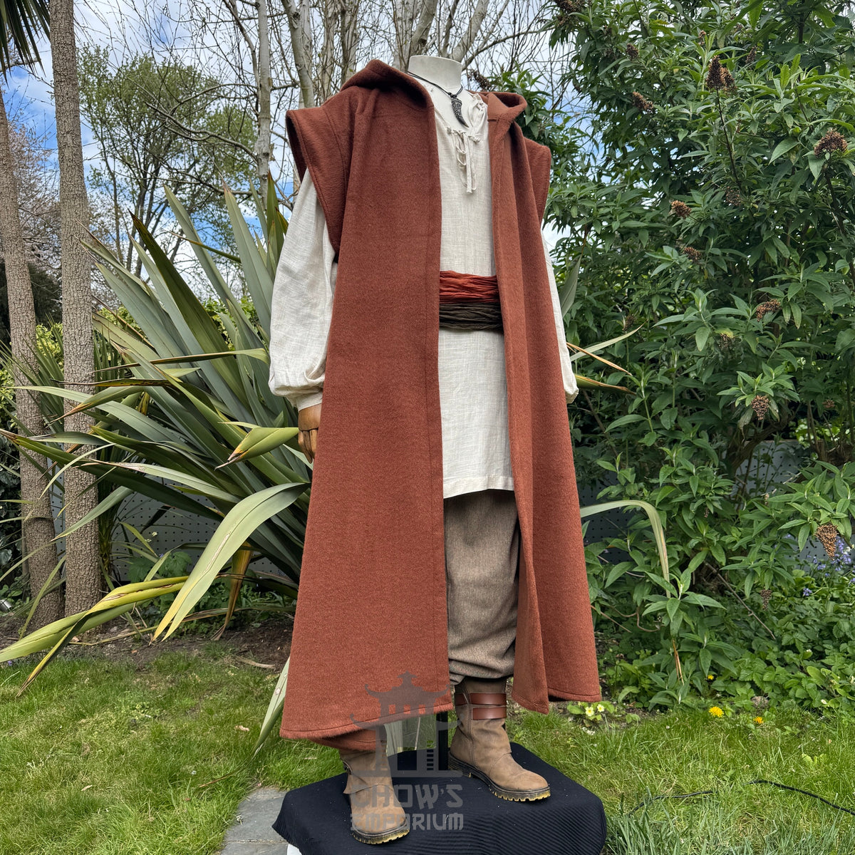 A mannequin wearing a medieval-style long rust hooded robe over a light beige shirt and brown trousers, standing side view with hood down outdoors in a garden with trees in the background. 