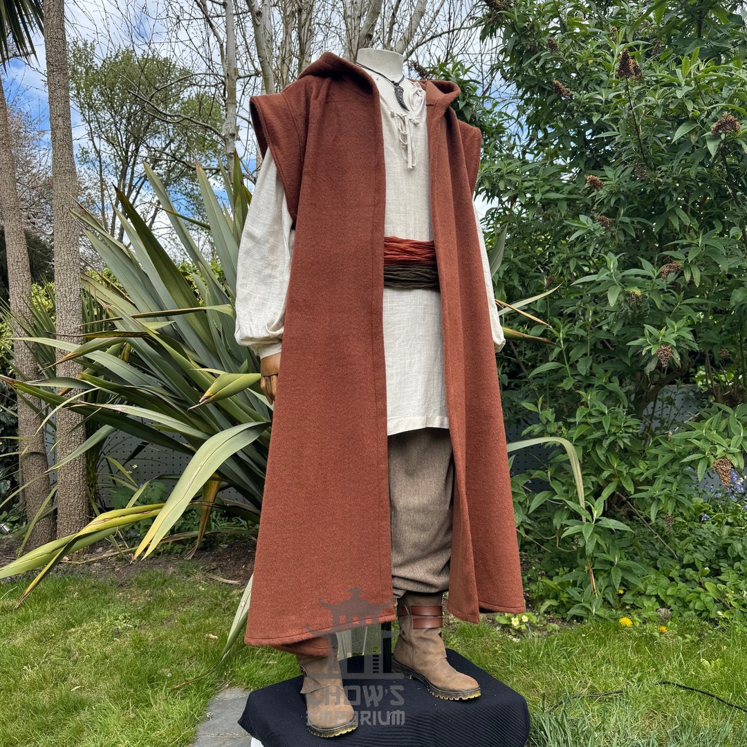 A mannequin wearing a medieval-style long rust hooded robe over a light beige shirt and brown trousers, standing side view with hood down outdoors in a garden with trees in the background. 