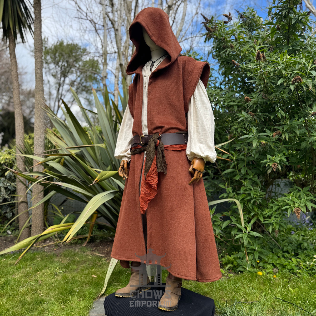 Mannequin wearing a medieval-style long rust robe with hood up, belted at the waist, showing sleeve from front side view, standing outdoors in a garden with trees in the background. 