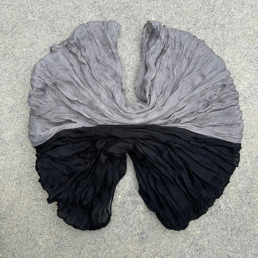 A lightweight, semi-sheer scarf with a horizontal color-blocked design, featuring a light gray top half and a black bottom half, laid flat on a textured gray concrete surface.