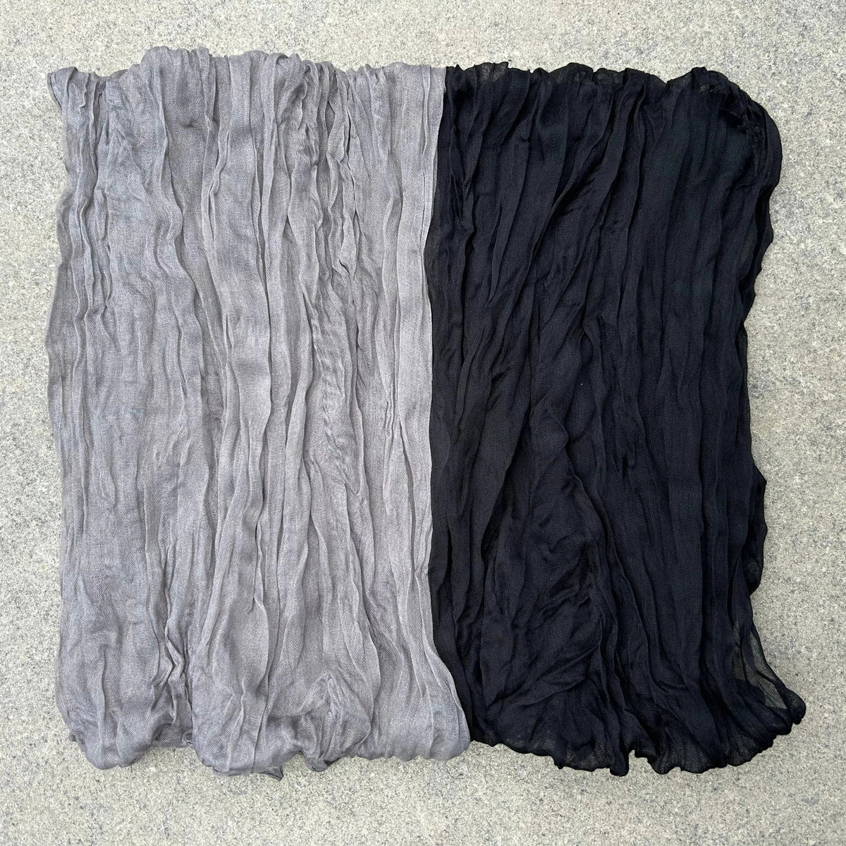 A lightweight, crinkled fabric scarf or shawl laid flat on a textured gray surface, featuring a horizontal color-blocked design with a light gray top half and a black bottom half.