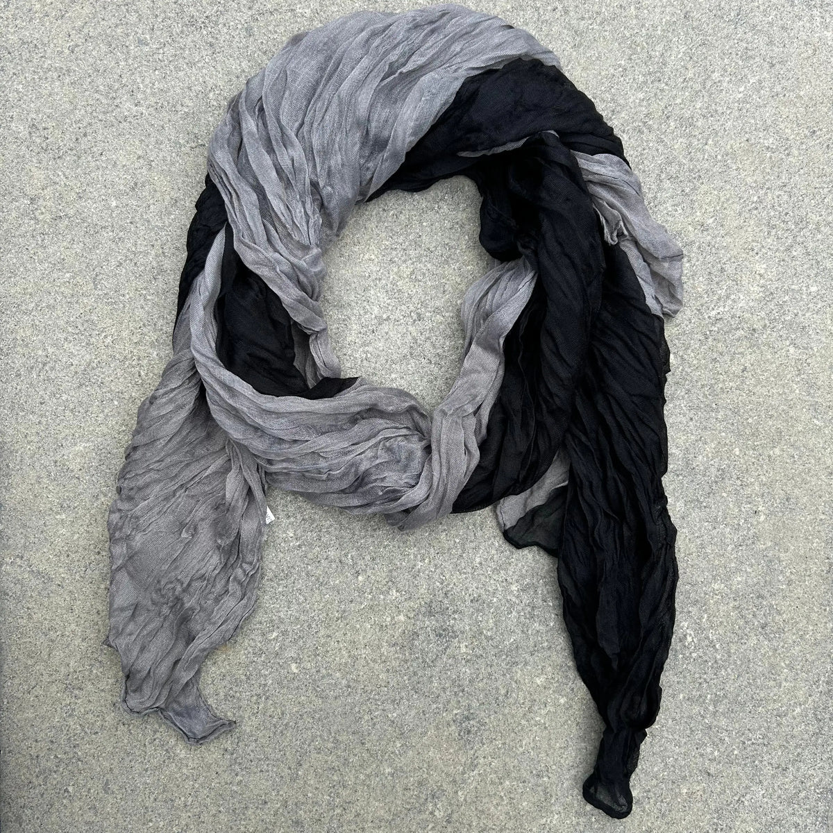 A lightweight crinkled scarf in a two-tone gray and black color-blocked design, laid flat on a textured gray concrete surface.