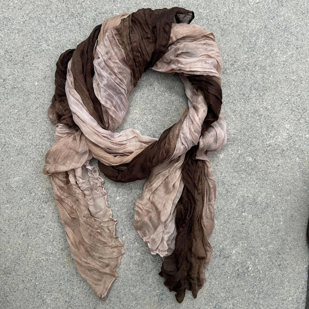 A lightweight, semi-sheer scarf with a shibori tie-dye pattern in shades of brown, beige, arranged in a loose loop on a textured gray surface.