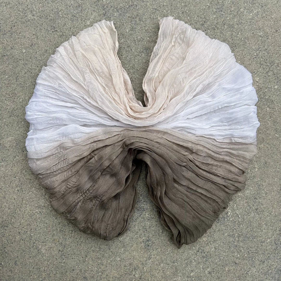 A lightweight, semi-sheer fabric scarf or shawl with a vertical ombré gradient, laid flat on a textured gray concrete surface. The top portion is off-white/cream, transitioning down through light taupe to a darker brown at the bottom.