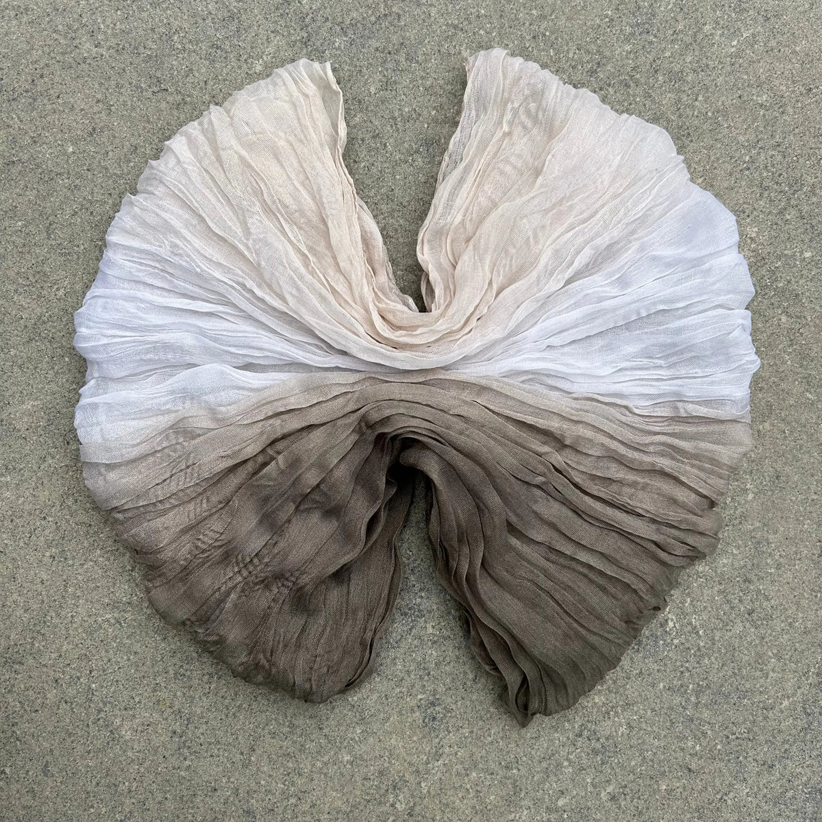 A lightweight, semi-sheer fabric scarf or shawl with a vertical ombré gradient, laid flat on a textured gray concrete surface. The top portion is off-white/cream, transitioning down through light taupe to a darker brown at the bottom.
