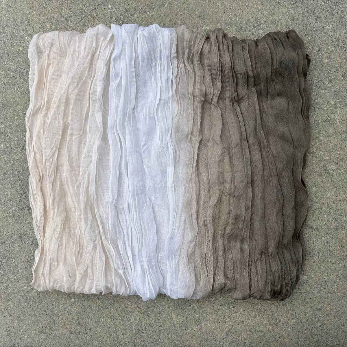 A square piece of sheer, crinkled fabric with a vertical ombré transition from light beige at the top, through white in the middle, to dark brown at the bottom, laid flat on a gray textured surface.