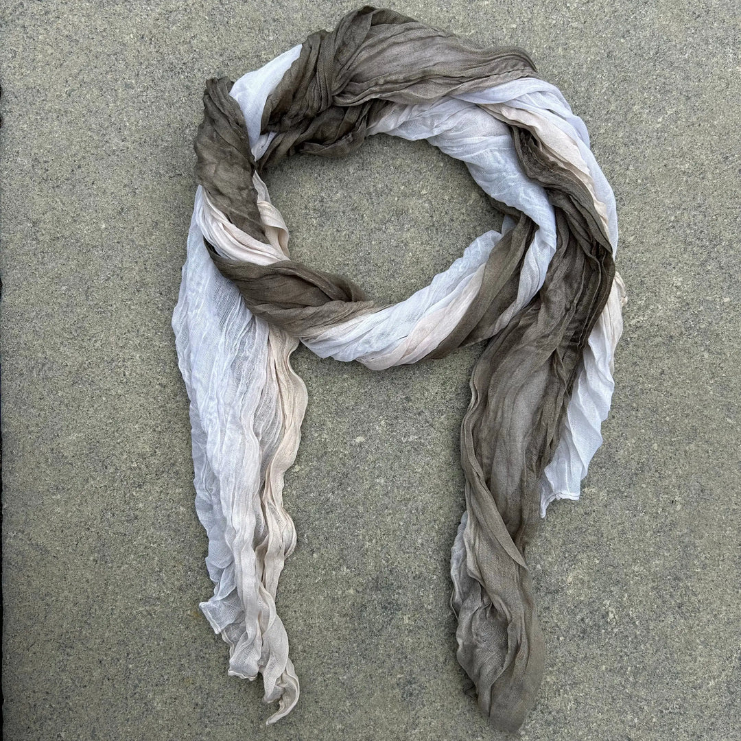 A lightweight, semi-sheer scarf with a gradient color transition from white to light beige and dark taupe-brown, arranged in a loose loop on a gray concrete surface.