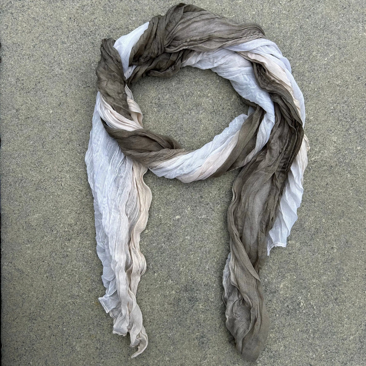 A lightweight, semi-sheer scarf with a gradient color transition from white to light beige and dark taupe-brown, arranged in a loose loop on a gray concrete surface.