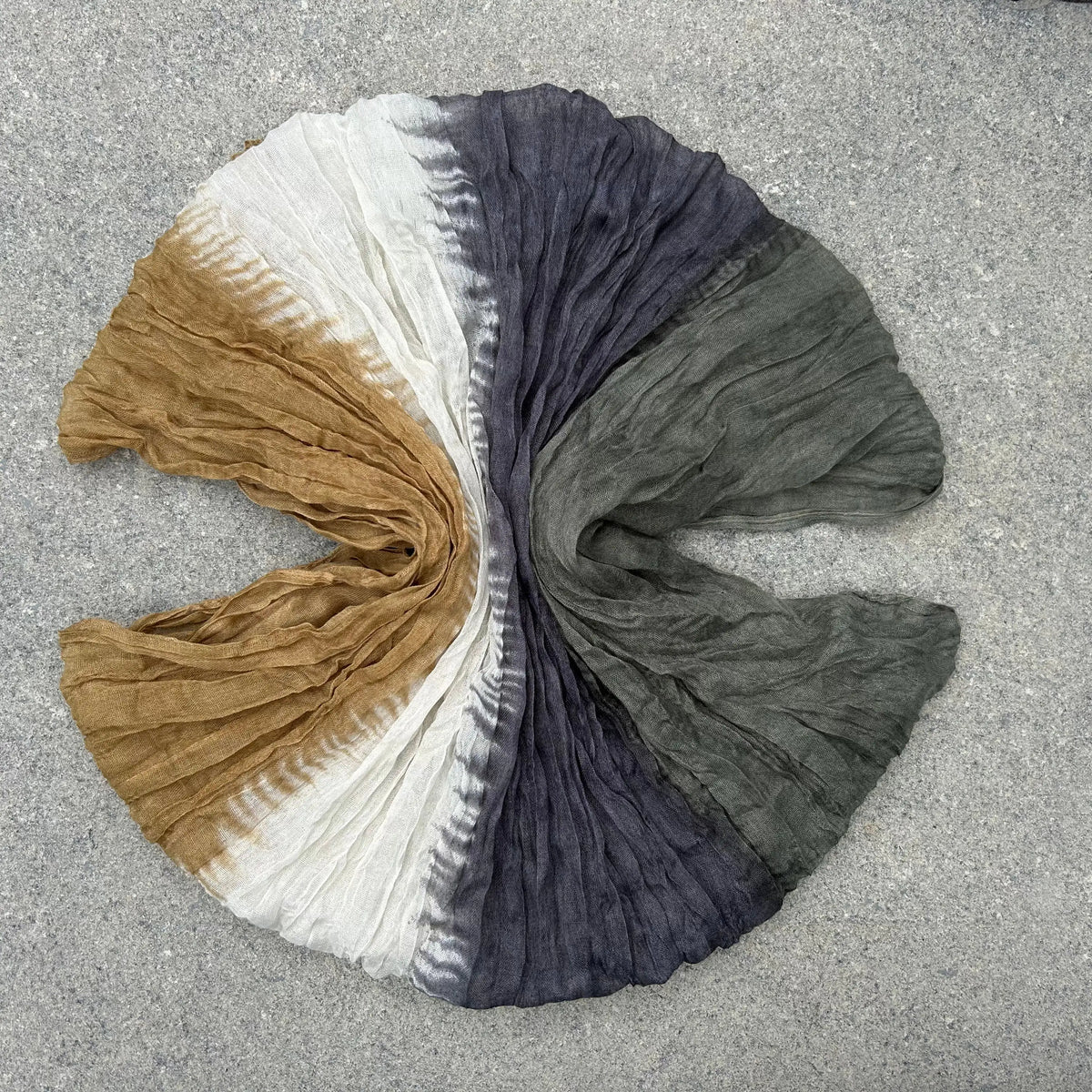 A crinkled fabric scarf or shawl laid flat on a gray textured surface, showing a tie-dye color gradient from tan and off-white on the left, through dark blue-gray in the center, to muted olive green on the right.