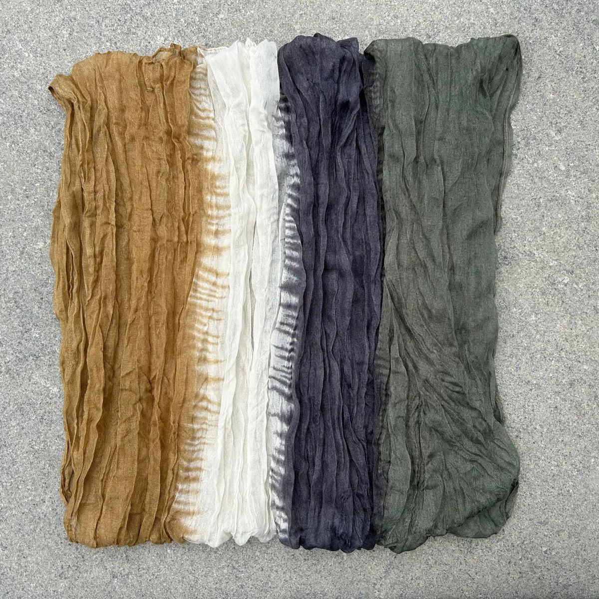 A lightweight crinkled scarf with a tie-dye pattern in charcoal gray, olive green, white, and tan, folded on a gray concrete surface.