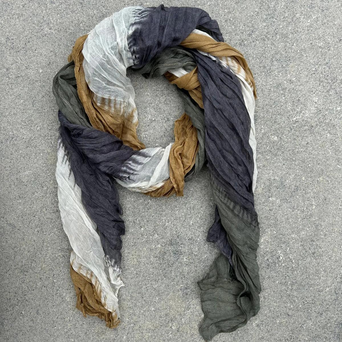 A lightweight crinkled scarf with a tie-dye pattern in charcoal gray, olive green, white, and tan, draped in a loose loop on a gray concrete surface.