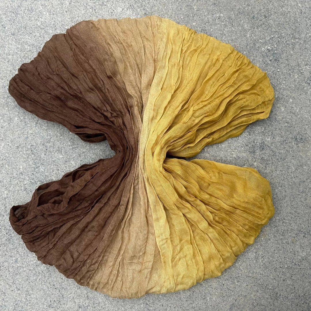 A crinkled fabric scarf or shawl laid flat on a gray textured surface, featuring a vertical ombré color transition from dark brown at the top, through tan, to mustard yellow at the bottom.