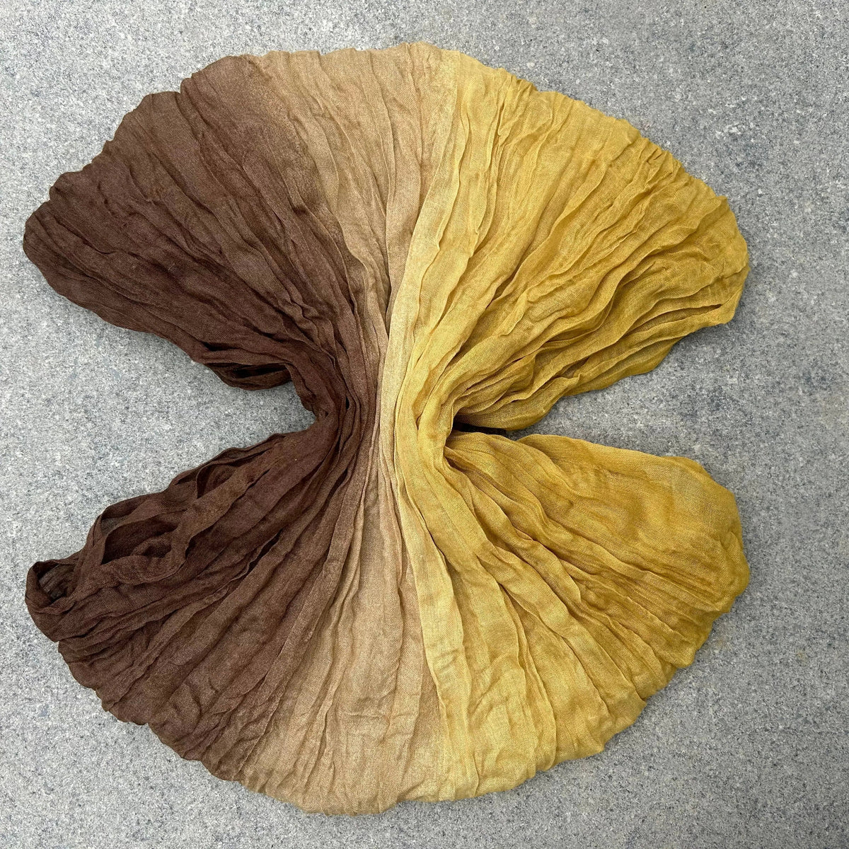 A crinkled fabric scarf or shawl laid flat on a gray textured surface, featuring a vertical ombré color transition from dark brown at the top, through tan, to mustard yellow at the bottom.