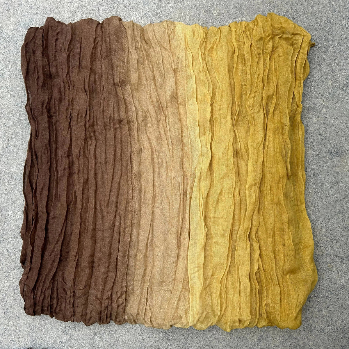 A square piece of crinkled fabric with a vertical ombre gradient transitioning from dark brown at the top, through tan, to mustard yellow at the bottom, laid flat on a gray textured surface.