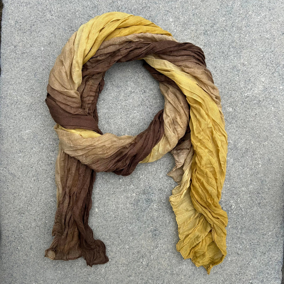 A crinkled scarf with a gradient color transition from dark brown to tan and mustard yellow, arranged in a loop on a gray textured background.