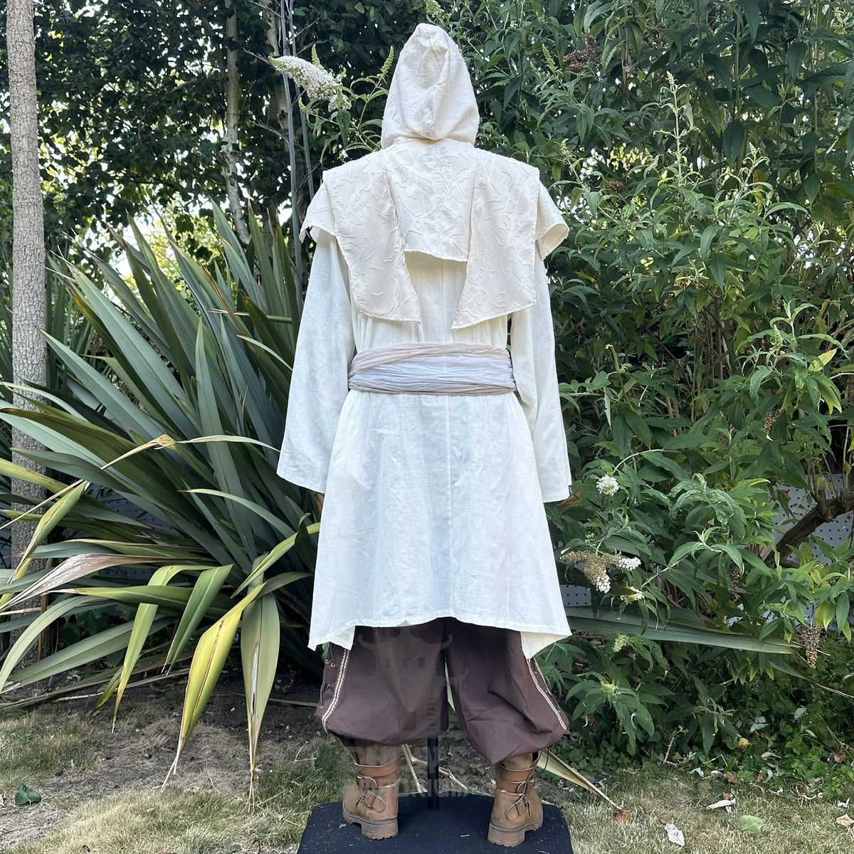 A costume or robe displayed on a mannequin outdoors, shown from the back. The outfit includes a cream hooded tunic with layered ruffle details, a light gray sash, brown baggy pants, and brown boots.