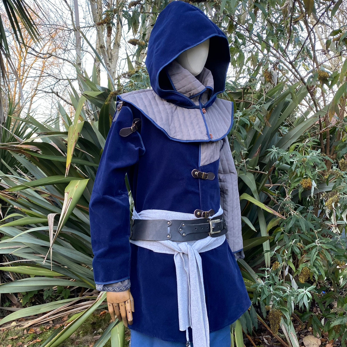 Mannequin in blue-grey hood and mantle, side view, outdoors in a medieval setting.